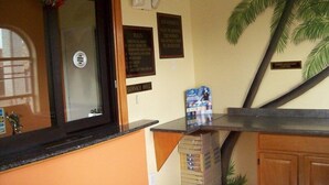 Un espace d’accueil comprenant un comptoir, une cloche pour signaler la présence d’un client, un panneau affichant les règles, fixé au mur, et une décoration en forme de palmier.