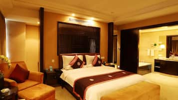 Quarto de hotel com uma cama grande, área de estar com poltrona e banquinho, escrivaninha com luminária e banheiro com espelho e pia.