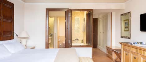 Habitación de hotel con cama, un banco, ventilador de techo, baño con bañera y un armario de madera.