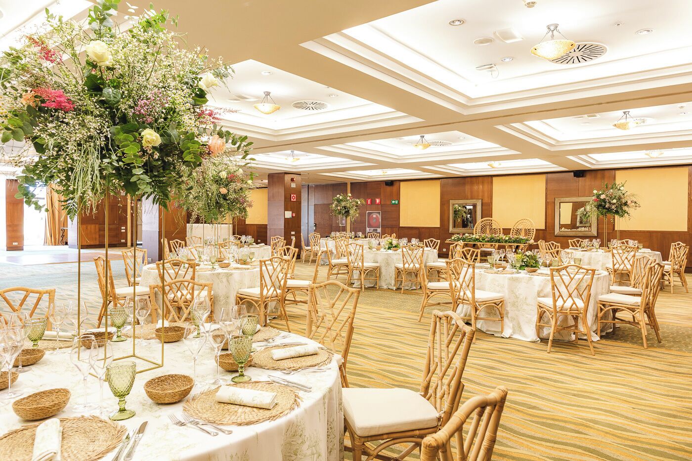 Un salón de banquetes con mesas redondas, sillas de mimbre y centros de mesa florales.