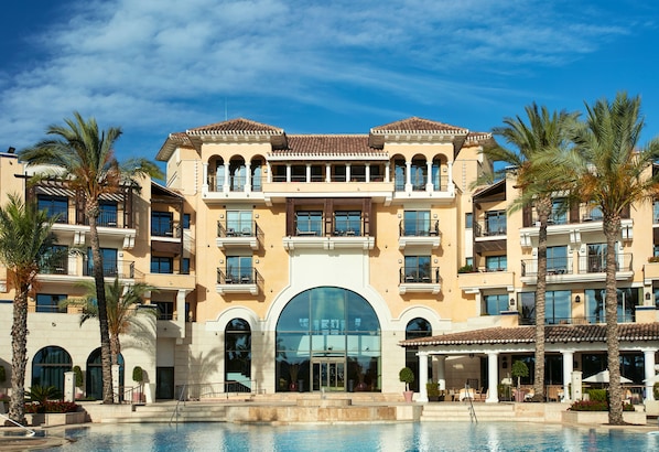 Un hotel de varios pisos con entrada central, balcones y zona de piscina.