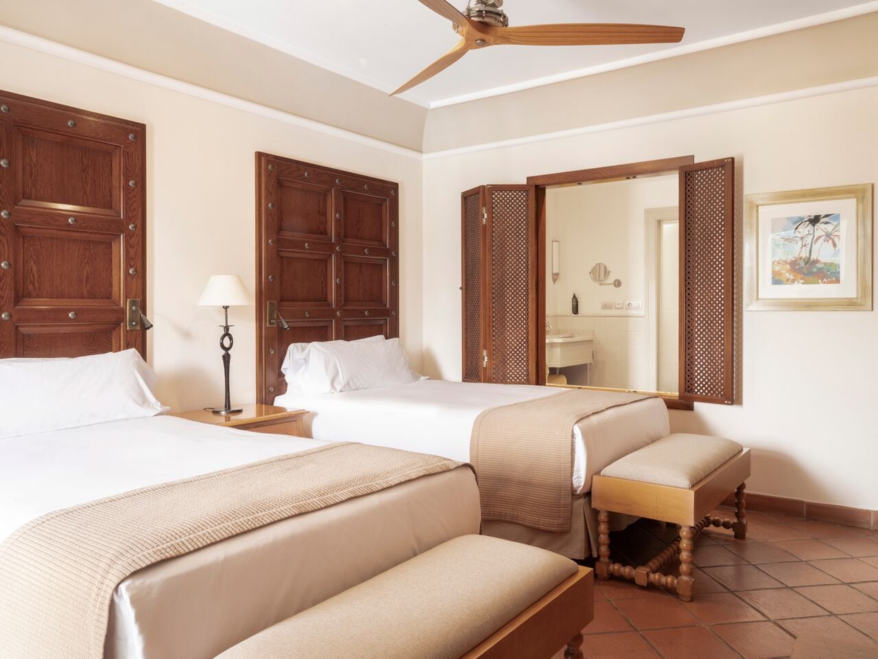 A hotel room with three beds, a ceiling fan, wooden doors, a bathroom mirror, and a framed picture on the wall.