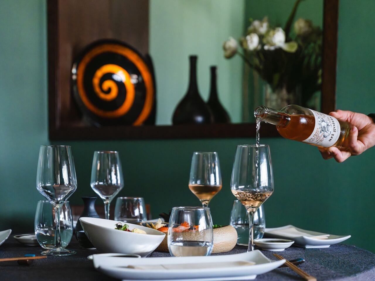 A dining table set with wine glasses, a bowl of food, and a bottle being poured.