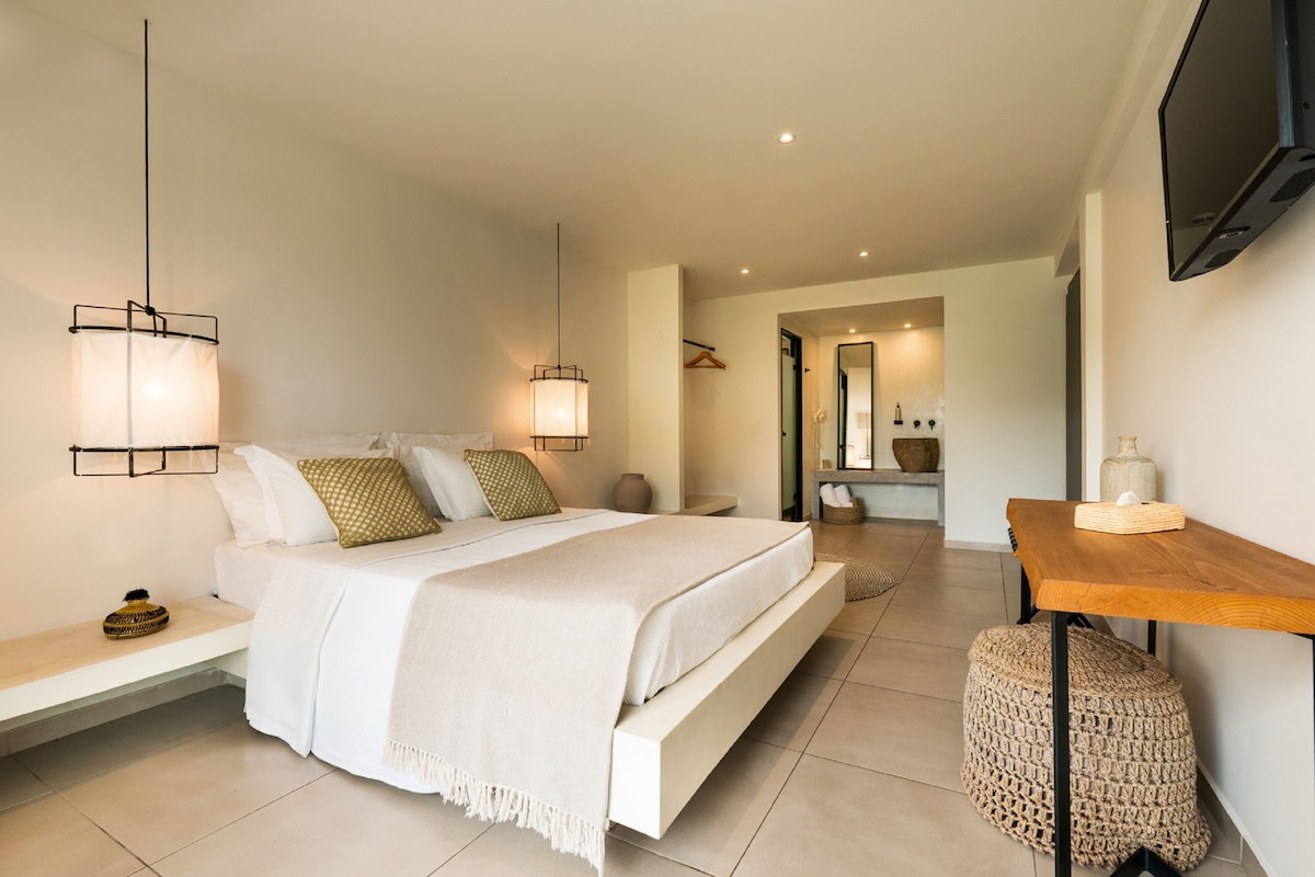 A modern bedroom with a bed, bedside tables, a wooden nightstand, a woven pouf, and a TV mounted on the wall.