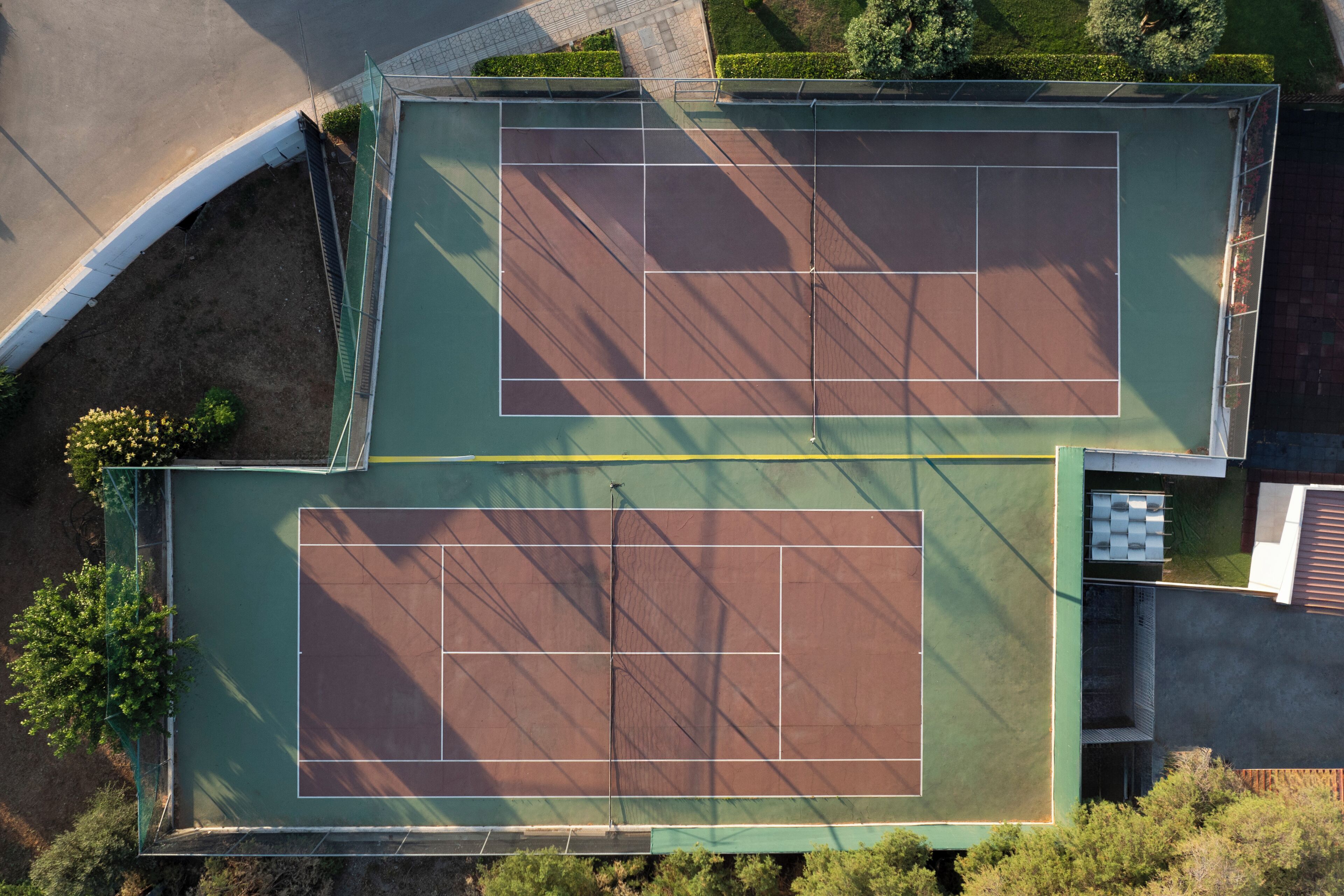 An aerial view of a tennis court complex with two courts, surrounded by greenery and adjacent to a road.