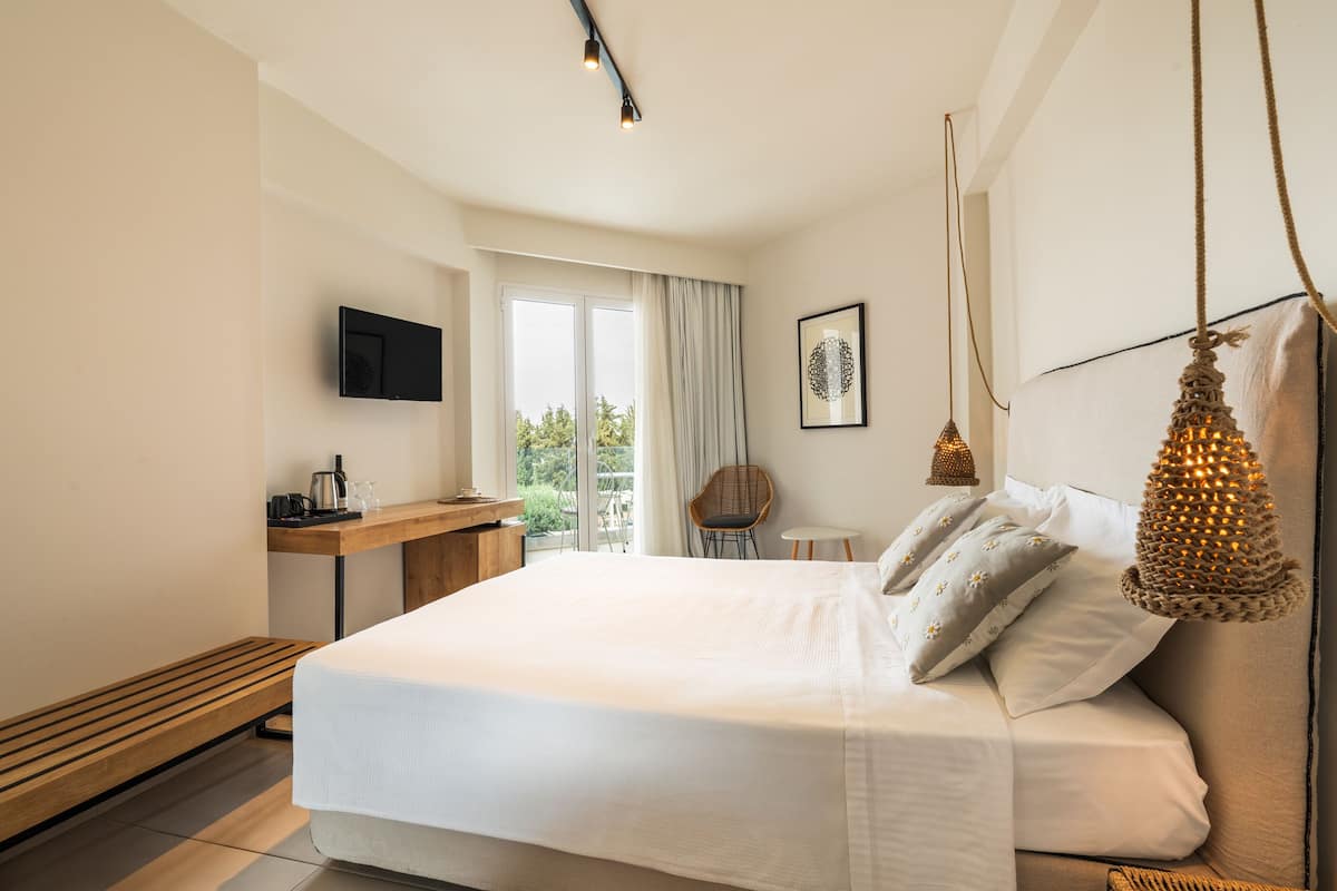 A modern hotel room with a large bed, a desk with a flat-screen TV, a window with curtains, and a hanging light fixture.