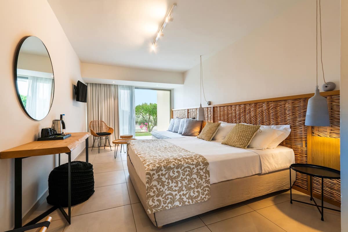 A modern hotel room with a large bed, a desk with a chair, a round mirror, and a view of greenery outside.
