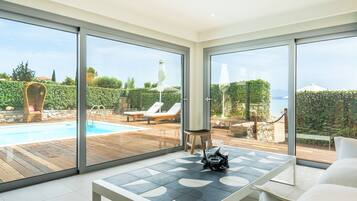 A modern living room with a large glass door leading to a pool area, a wooden deck, and a view of the sea.