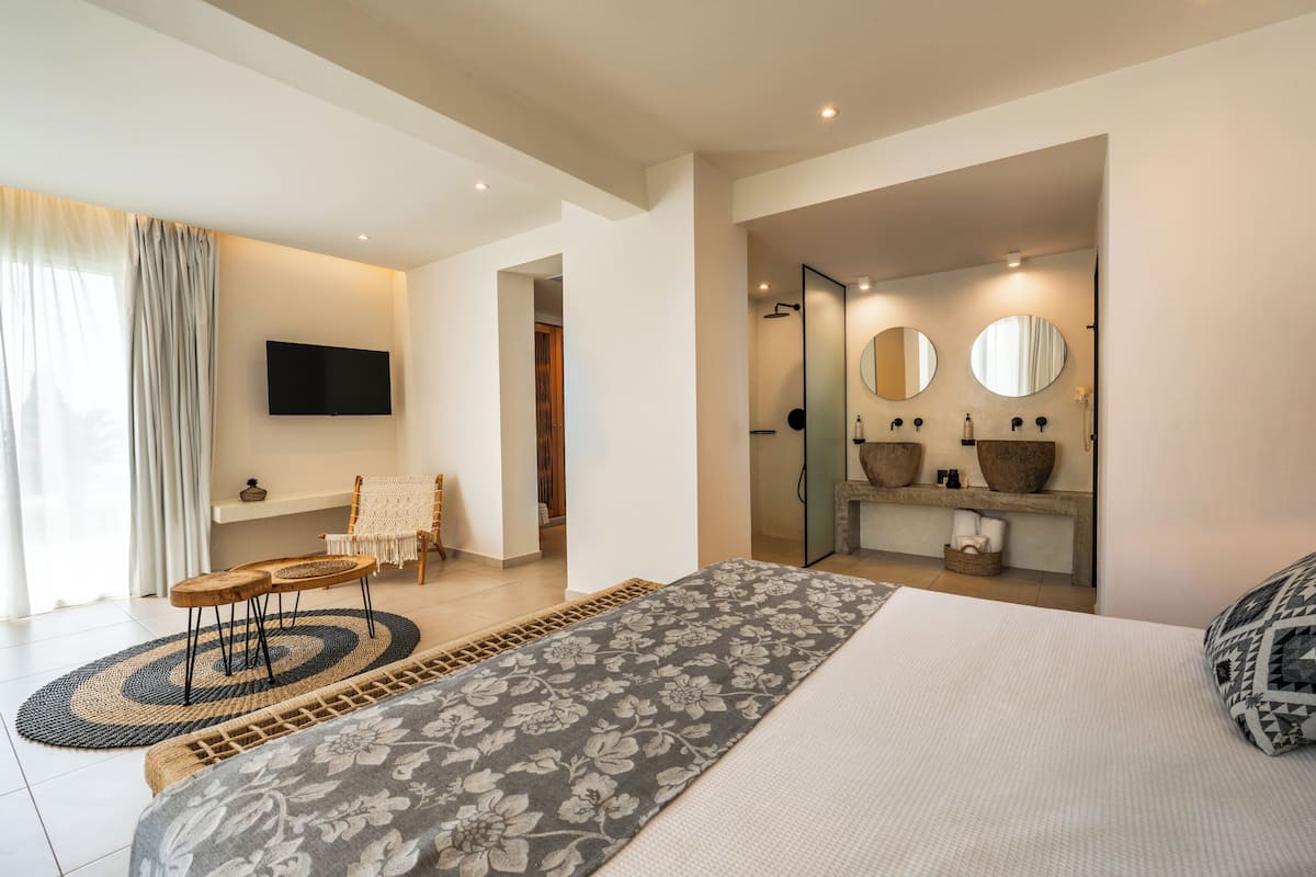 A modern bedroom with a large bed, a TV mounted on the wall, a small seating area, and a bathroom with a large mirror and sink.