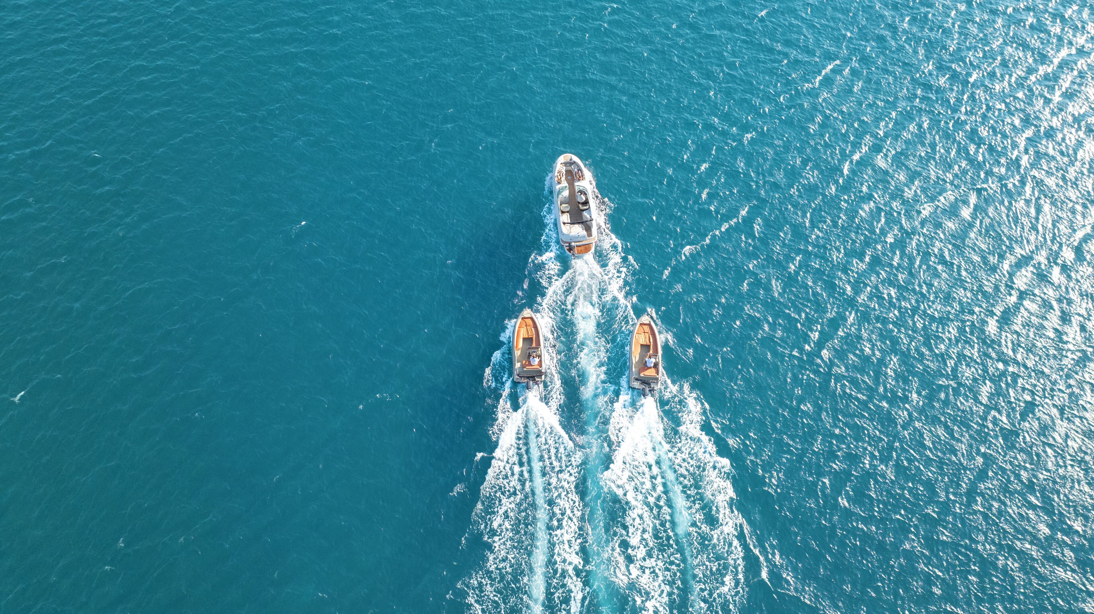 An aerial view of a yacht with two smaller boats following closely behind, creating a wake in the water.