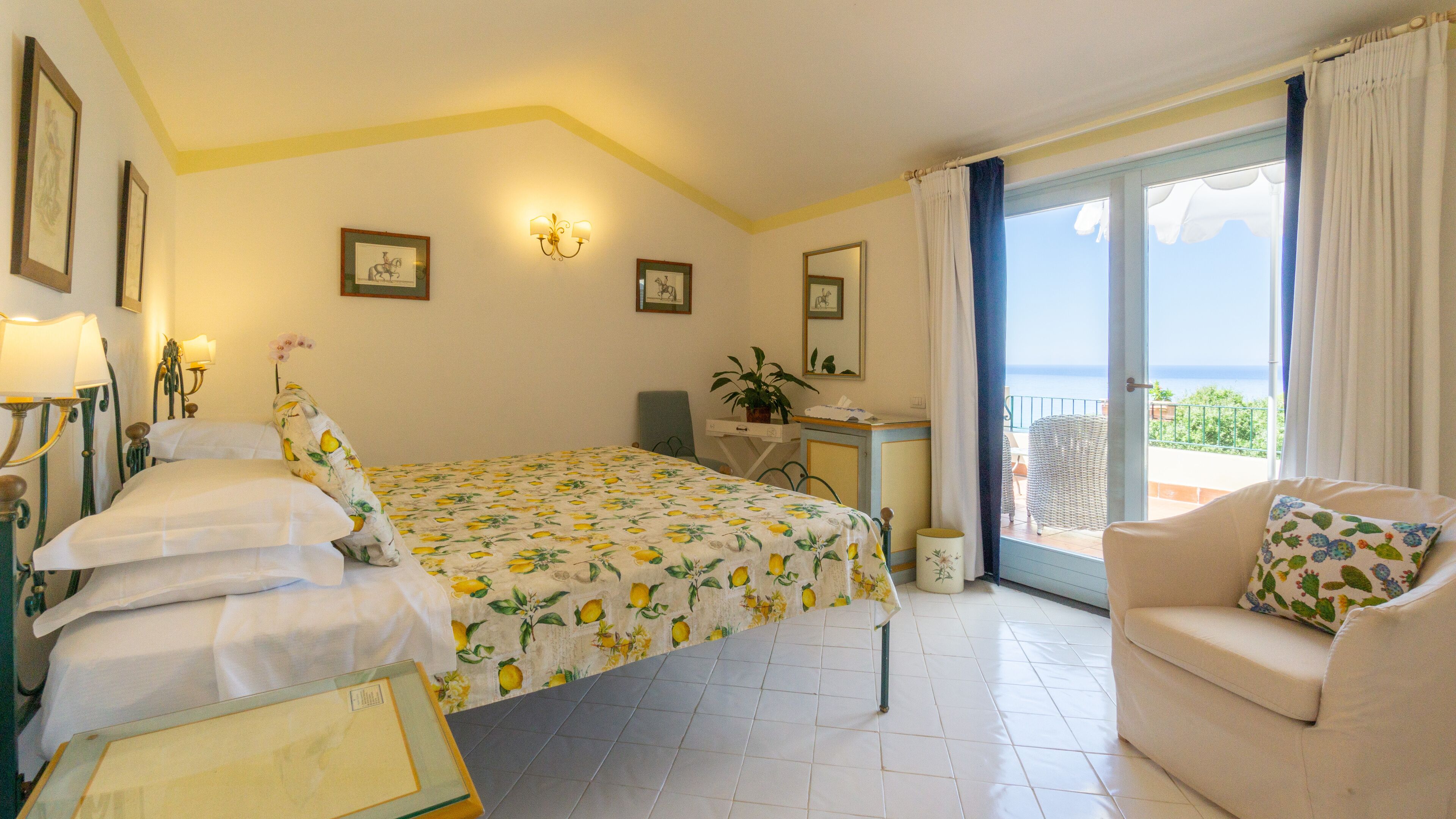 A hotel room with a bed, a desk, a chair, a balcony with a view, and a potted plant.