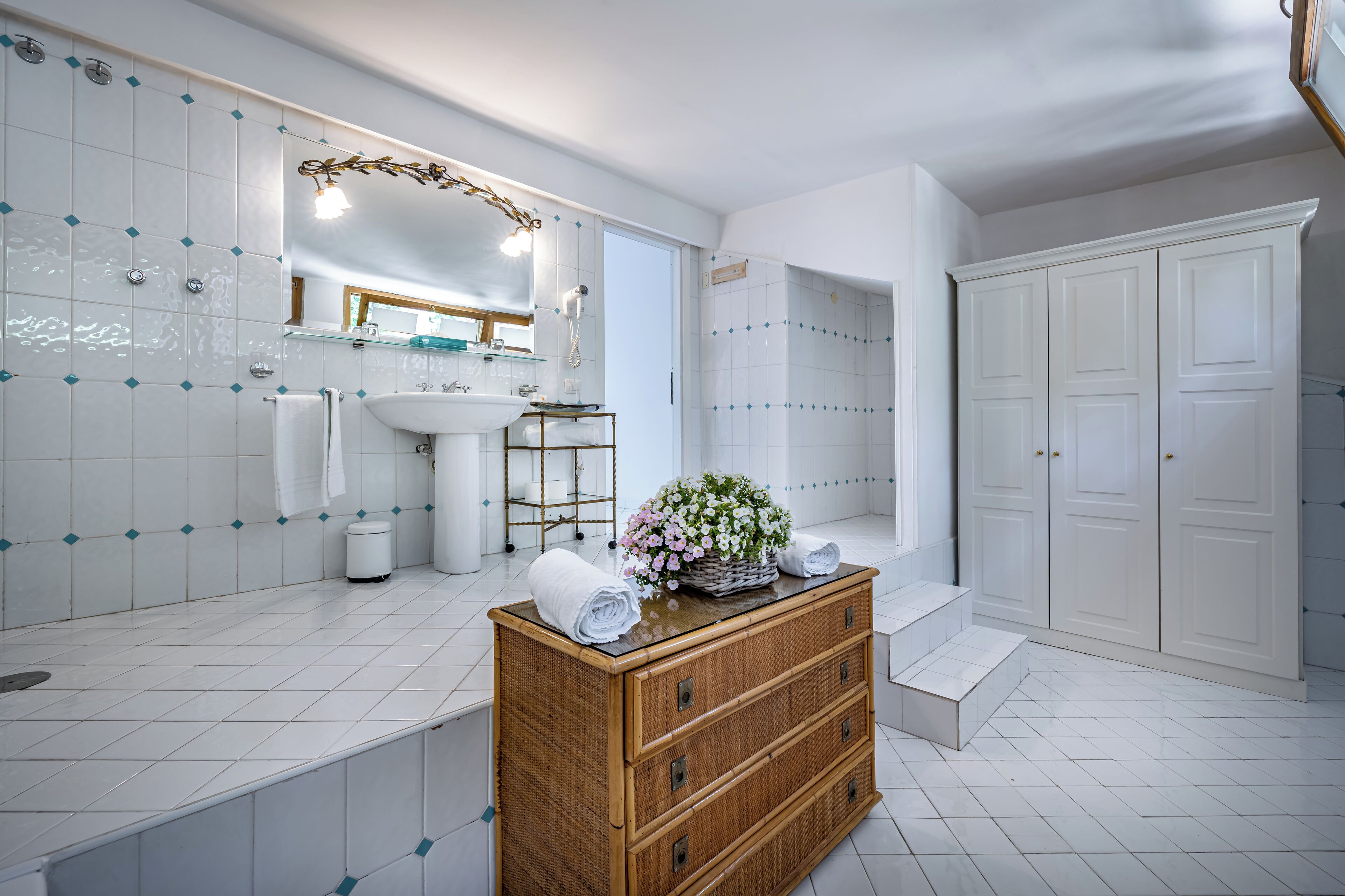 A modern bathroom with white tiles, a large mirror, a sink, and a wooden vanity with a basket of flowers.