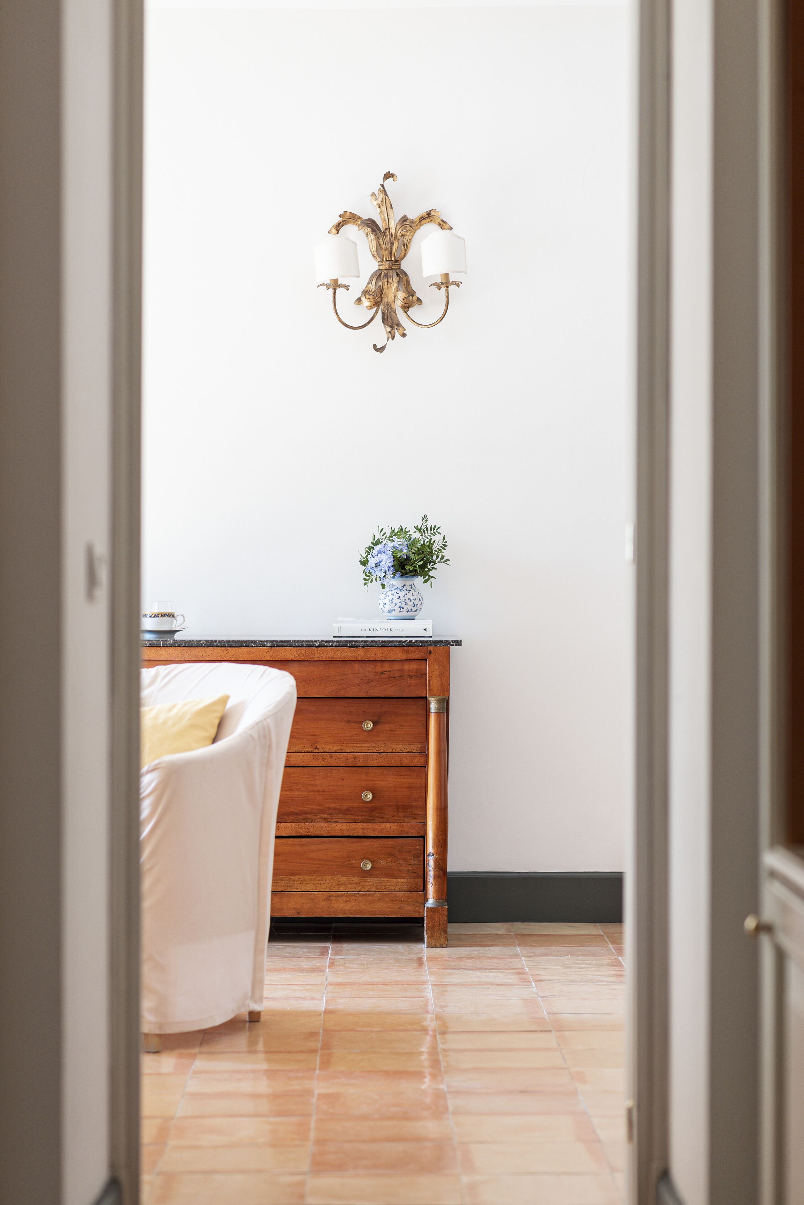 Un dormitorio con un aparador de madera, una silla blanca, un florero con flores y un aplique de pared.
