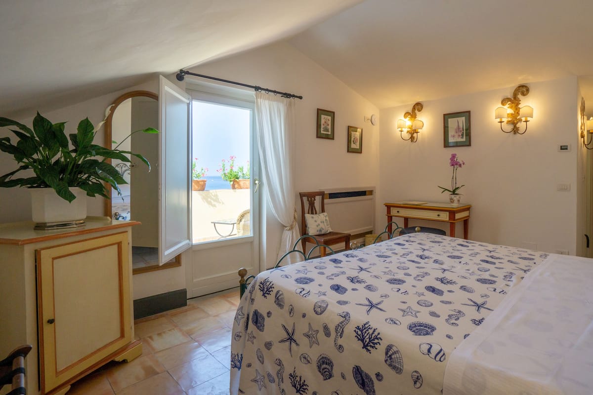 A bedroom with a bed, a desk, a chair, a potted plant, and a view of the outside.