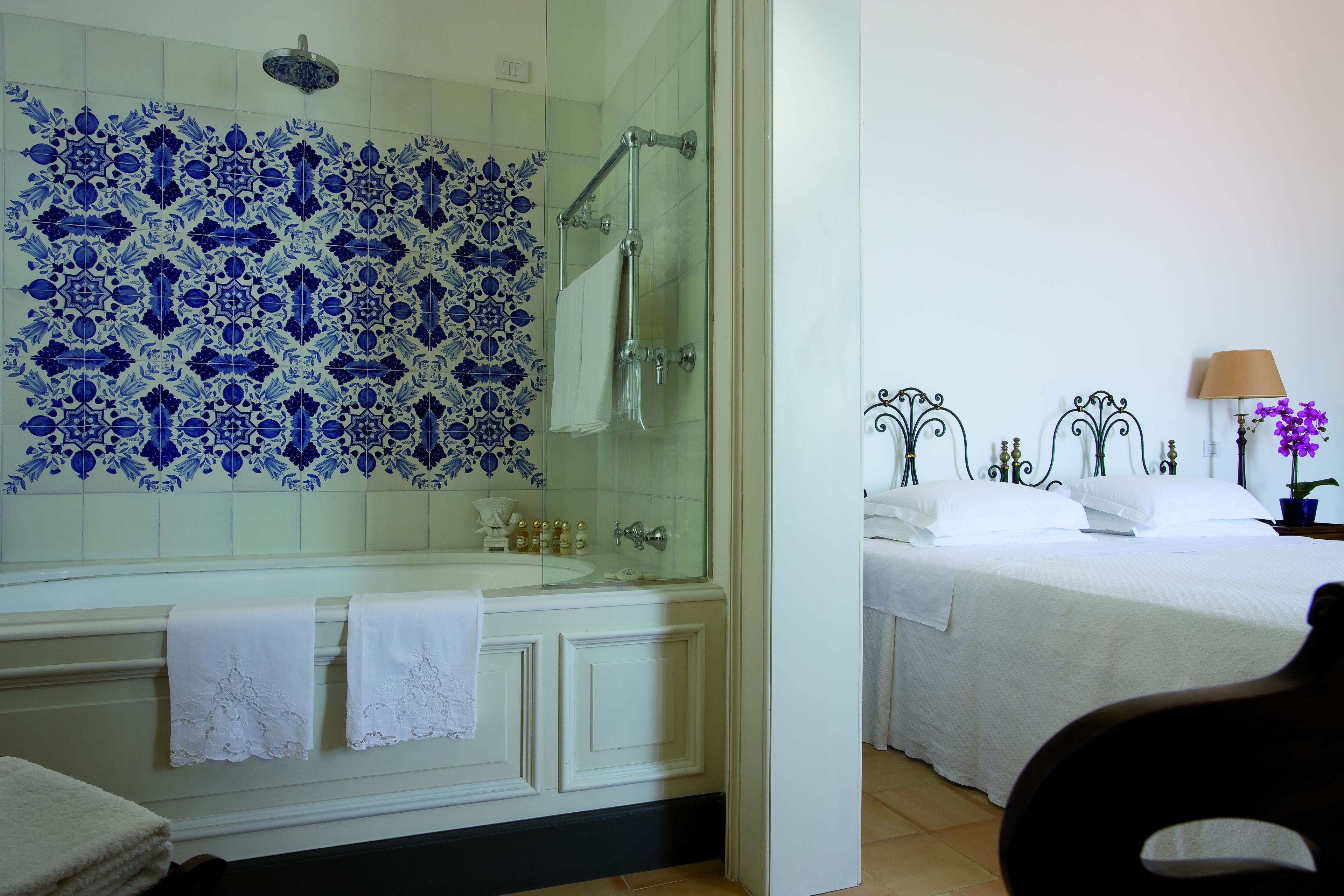 A bathroom with a glass-enclosed shower, a white bathtub with decorative tiles, and a bed with white bedding and a decorative headboard.