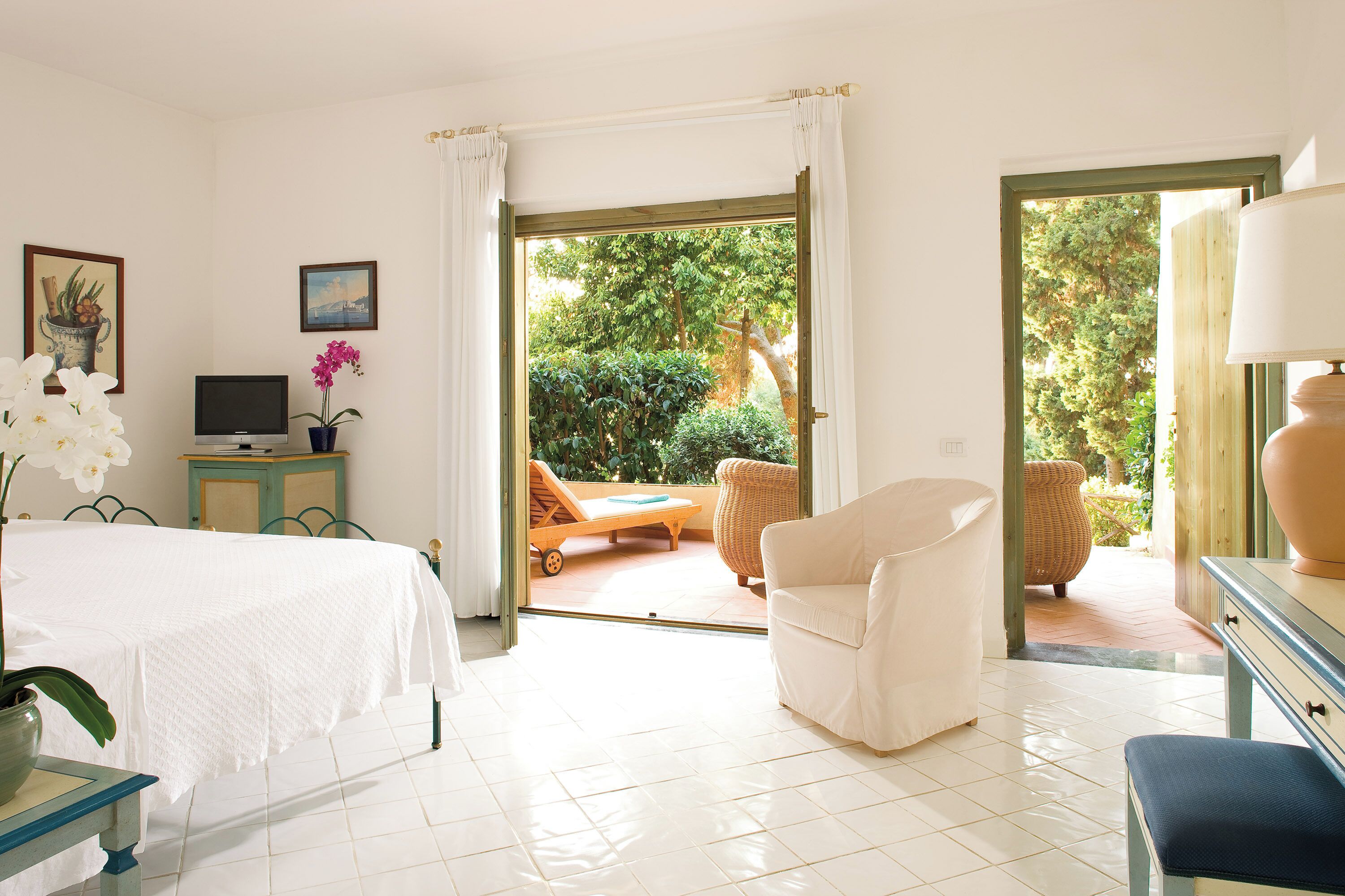 A hotel room with a bed, a TV, a desk with a lamp, a chair, and a view of the garden through the door.