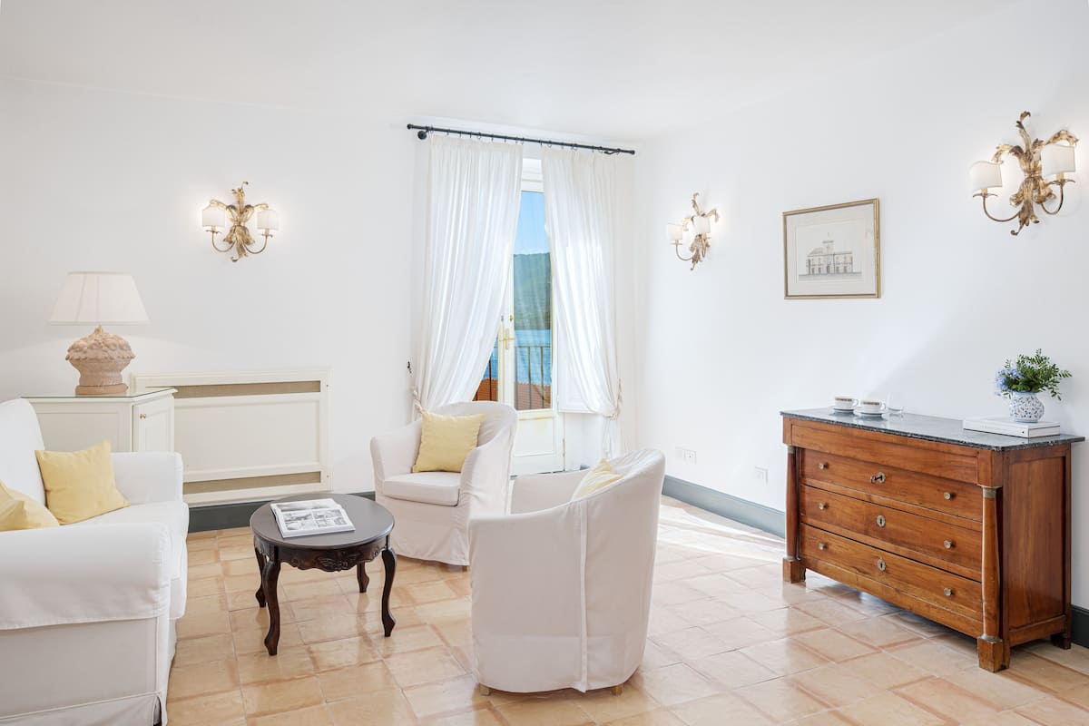 A living room with a white sofa, a white armchair, a wooden cabinet, and a window with white curtains.