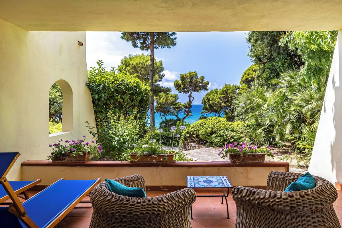 A terrace with wicker furniture, potted plants, and a view of the sea.