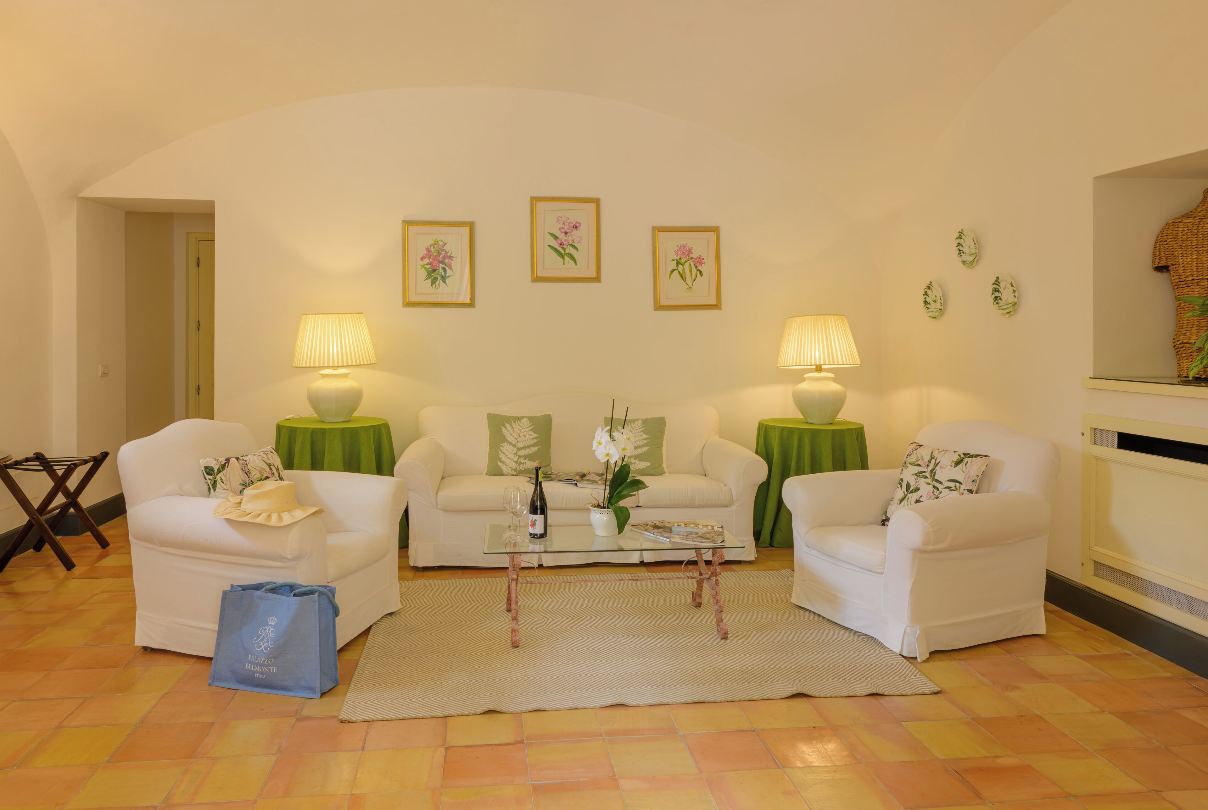 Sala de estar com dois sofás brancos, uma mesa de centro de vidro e duas luminárias. Quadros florais emoldurados na parede e toalha de mesa verde na mesa de centro.