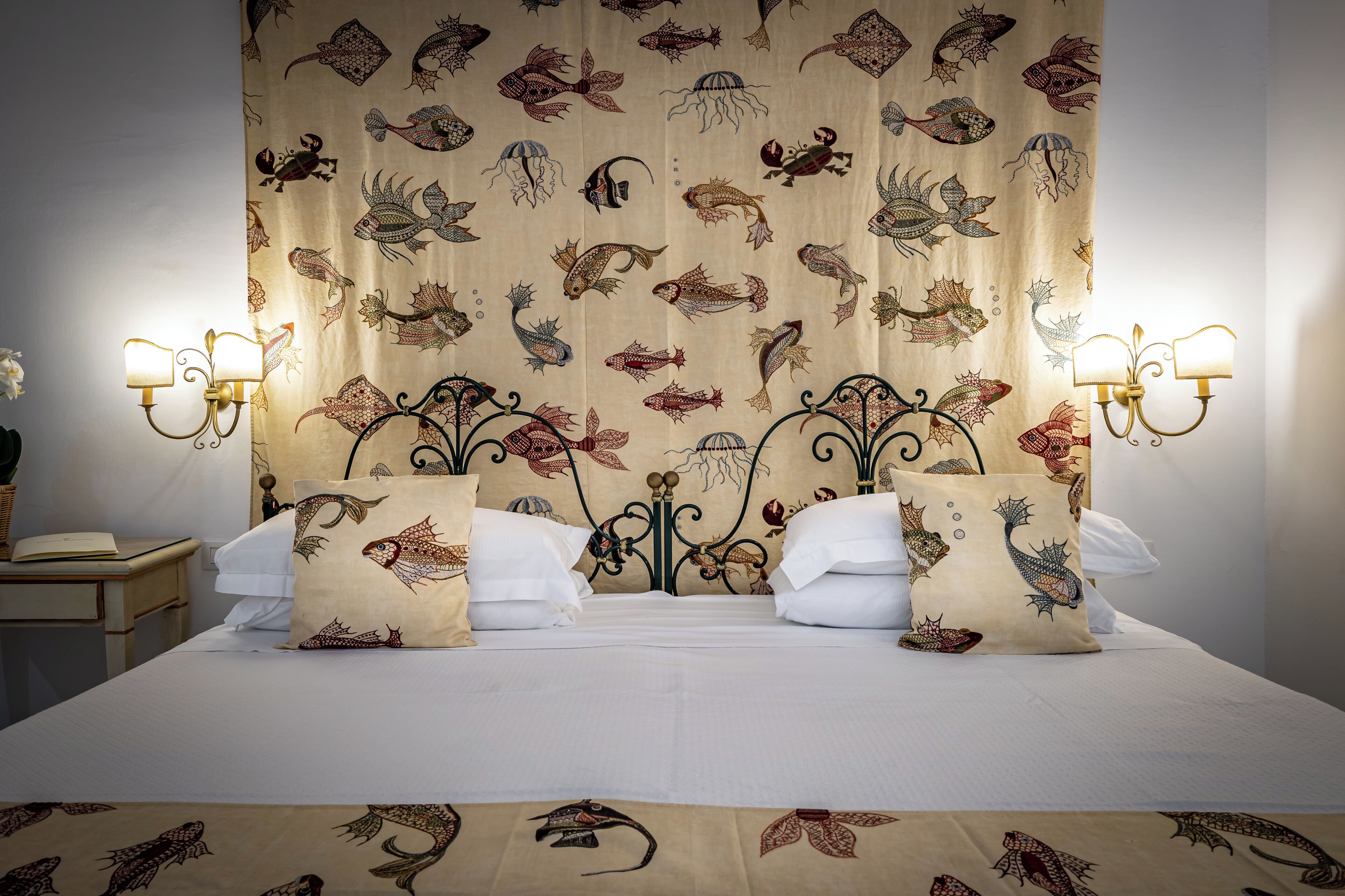 A bed with a headboard featuring a fish pattern, two wall-mounted light fixtures, and a bedside table with a book and a plant.