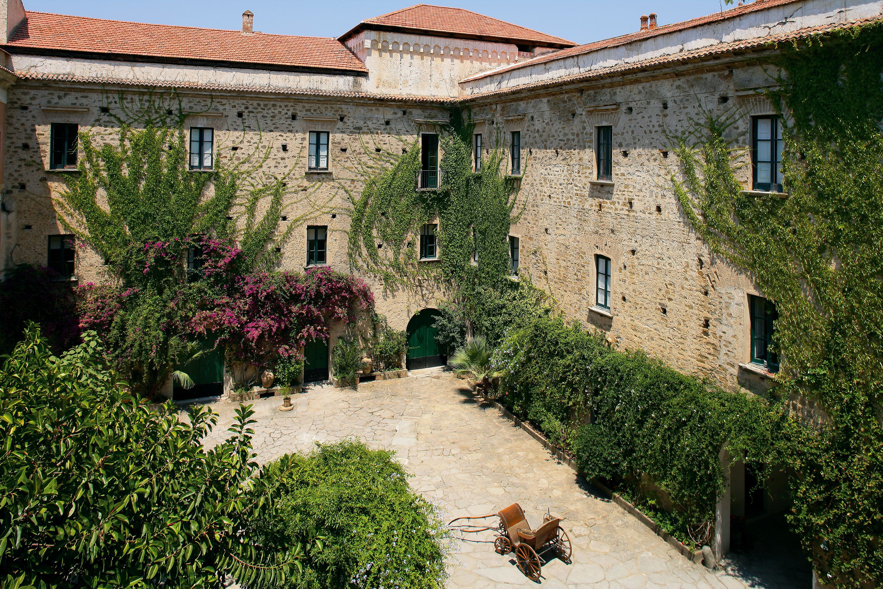 Un cortile con pavimento in pietra, una carrozza e vegetazione lussureggiante.