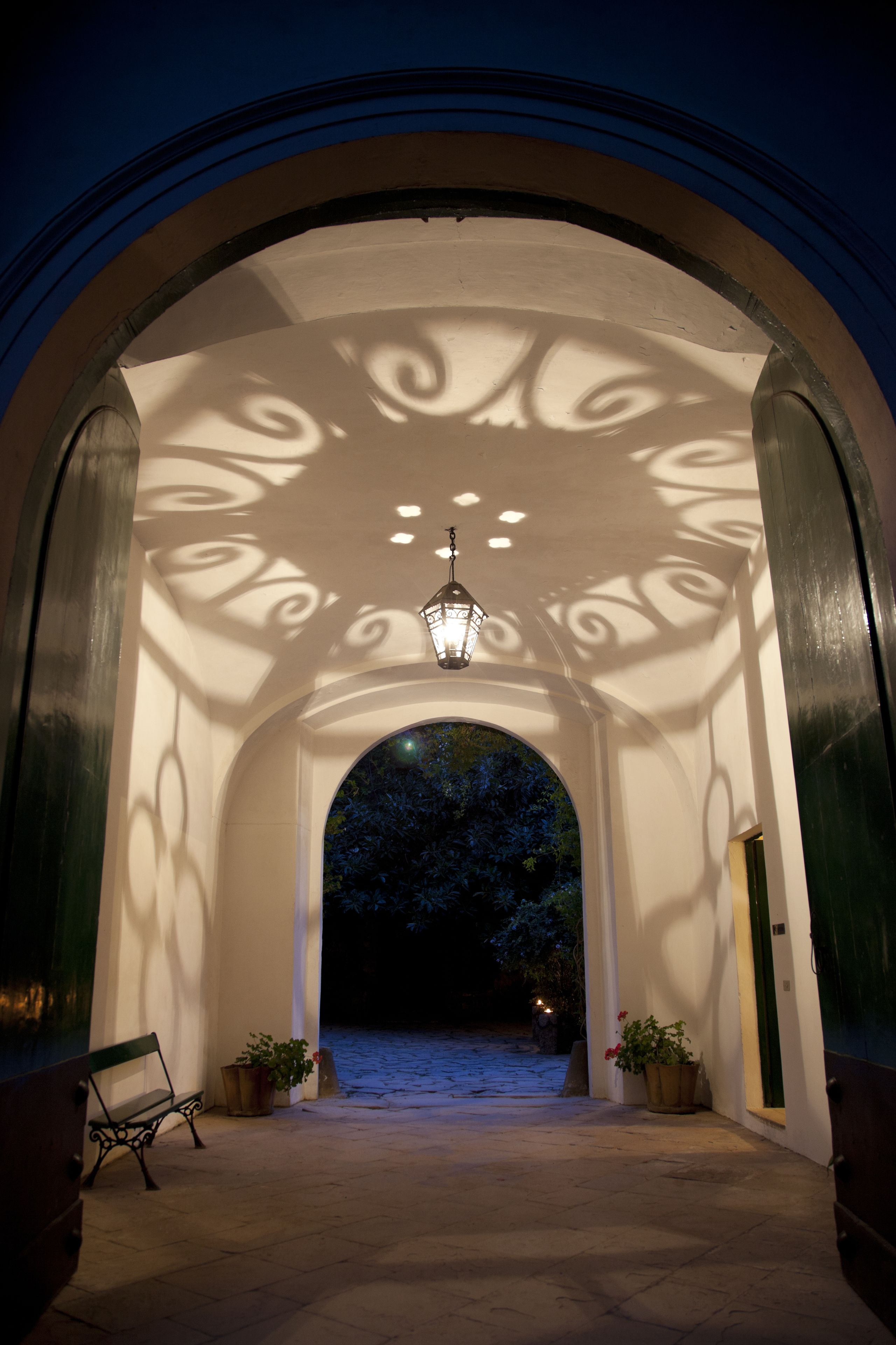 An arched entrance with a bench and potted plants, a hanging lantern, and a view into a dark area beyond.