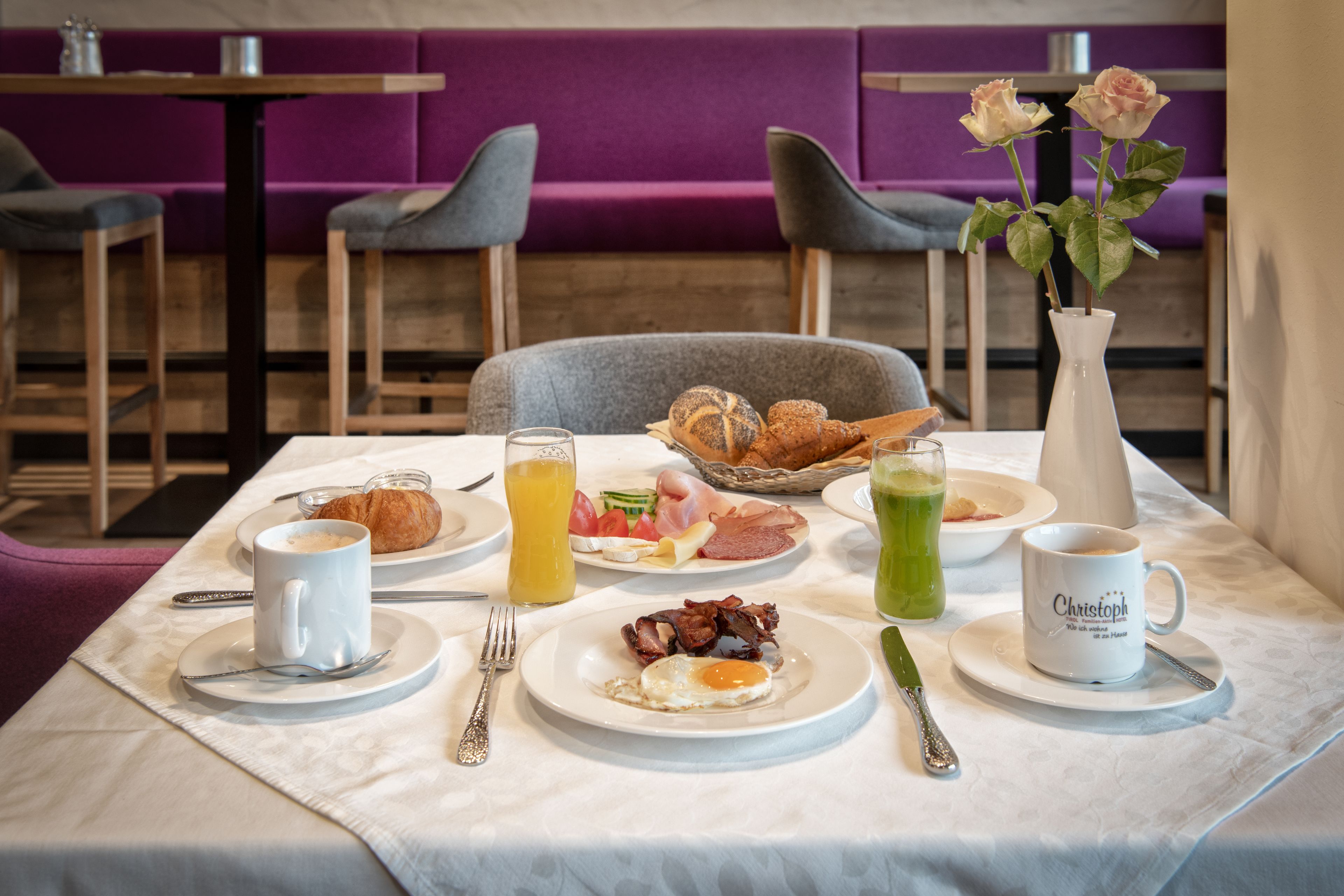 Mesa de desayuno preparada con croissants, huevos, tocino y jugo, en un comedor moderno con detalles morados.