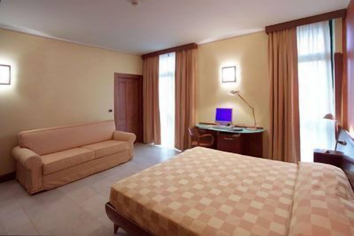 A hotel room with a bed, a sofa, a desk with a computer, and curtains.