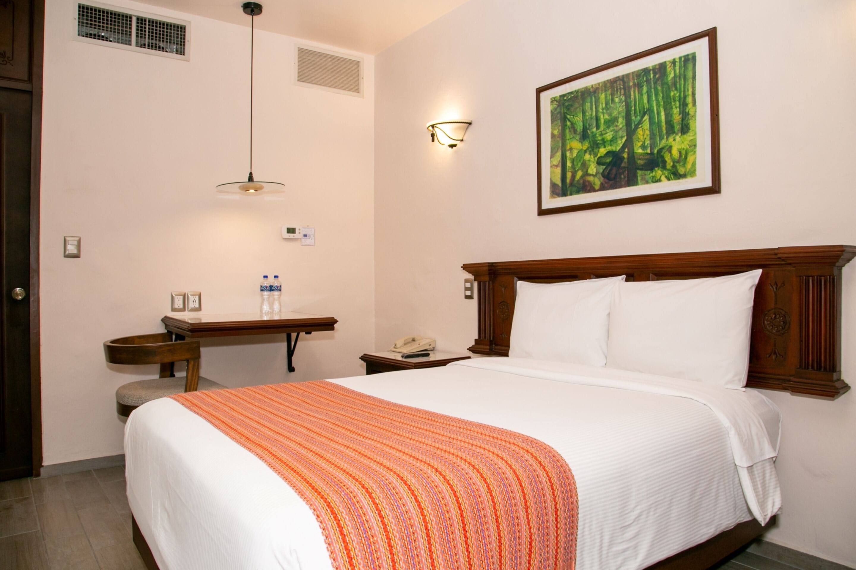 Une chambre d’hôtel avec un grand lit, un bureau avec un téléphone, une chaise, une lampe fixée au mur et un tableau représentant une forêt.