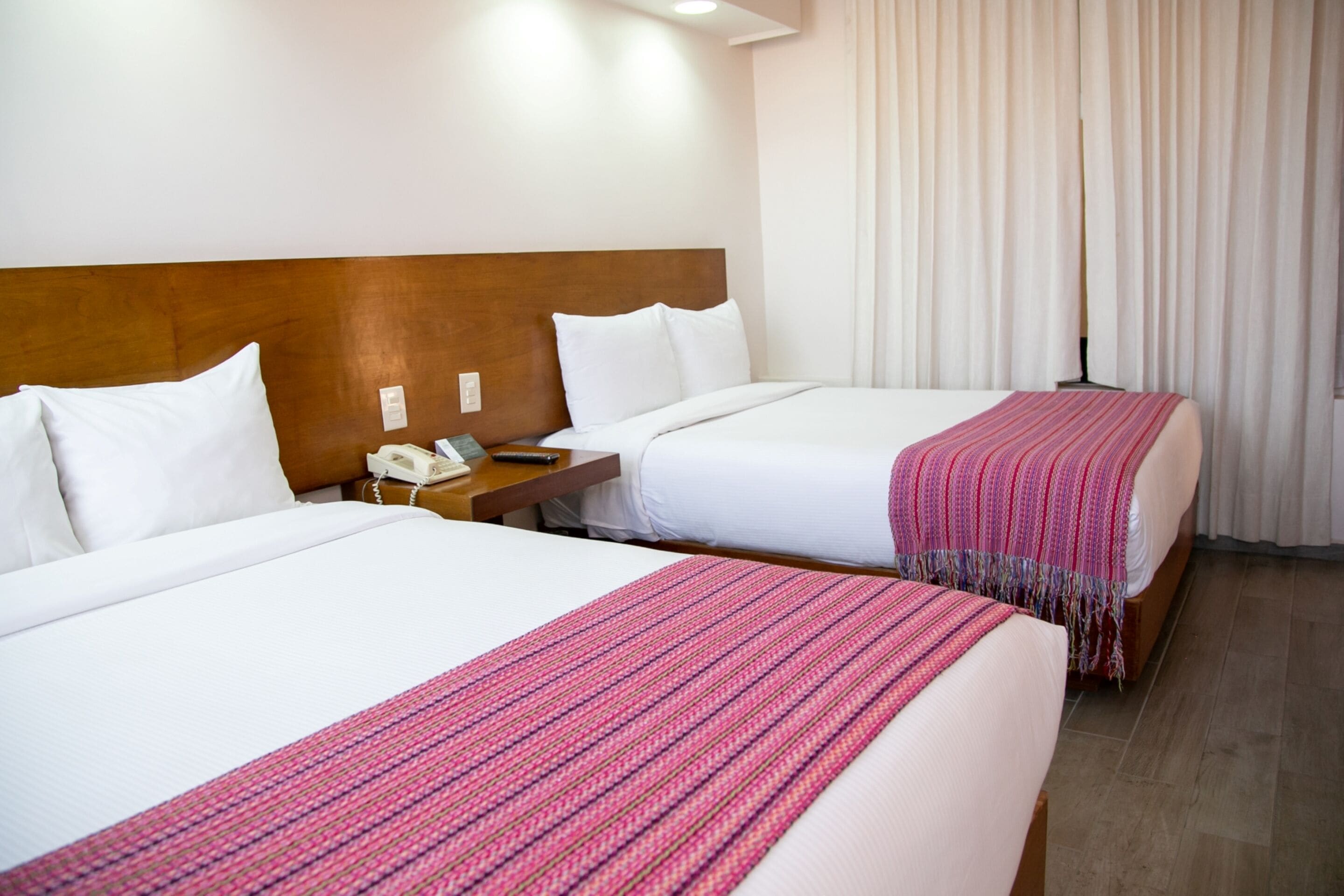A hotel room with two beds, a wooden headboard, white bedding, a telephone, and a window with curtains.