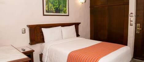 A hotel room with a wooden bed, white linens, a wooden nightstand, a desk, and a framed painting on the wall.