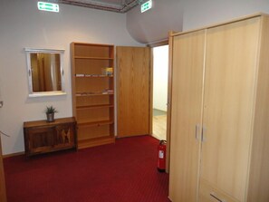 Ein Zimmer mit rotem Teppich, einem Holzschrank, offenen Regalen, einem kleinen Schrank, einer Topfpflanze und einem Feuerlöscher.