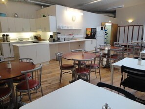 A dining area with round tables and metal chairs, a kitchen in the background, and a white color scheme.
