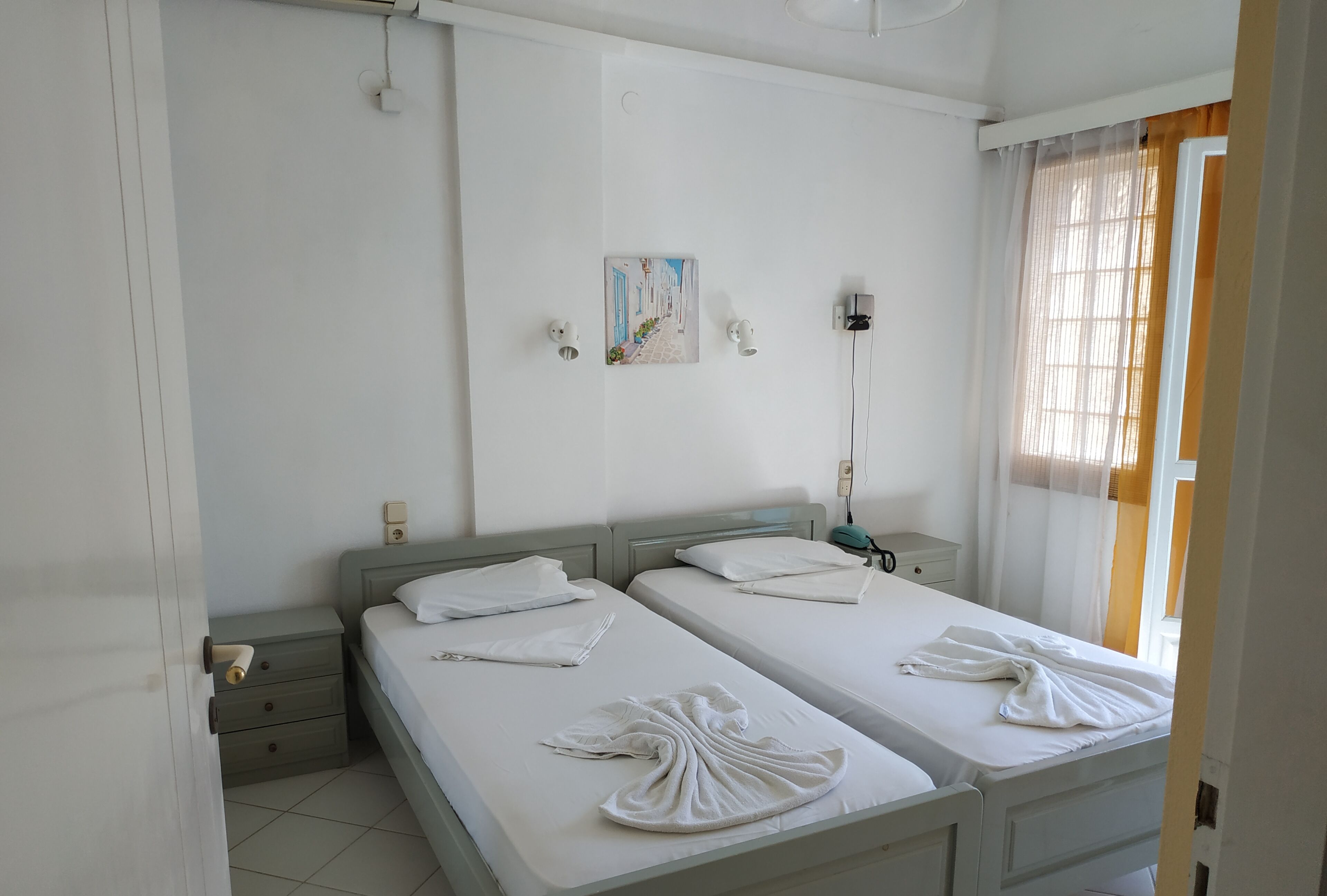 A hotel room with two beds, a painting on the wall, a bedside table with a lamp, and a window with curtains.