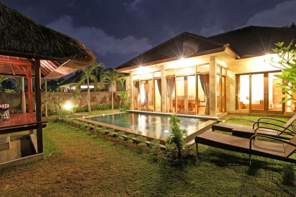 A well-lit outdoor pool area with a house, a thatched-roof structure, and a chair.