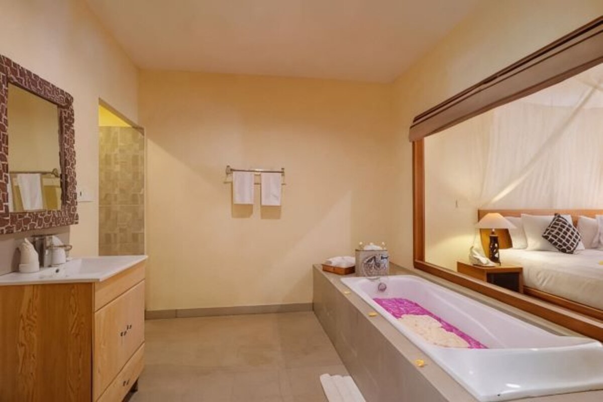 A hotel bathroom with a bathtub, a vanity with a mirror, and a bed visible through a large window.