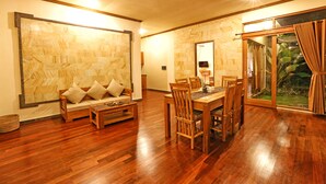 A spacious living area with a wooden floor, a sofa with cushions, a dining table with chairs, and a large window with curtains. - Bhanuswari Resort & Spa (Ubud)