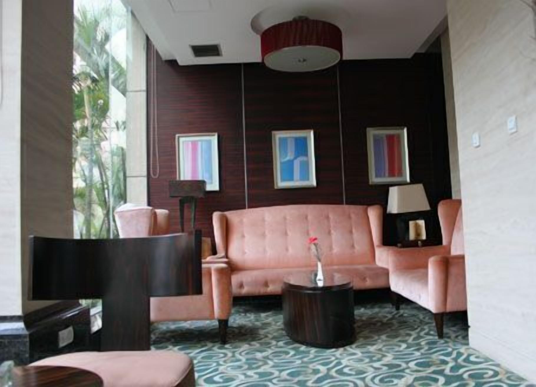 A modern lounge area with a pink sofa, a round coffee table, and a dark armchair. There are framed artworks on the wall and a lamp on a side table.