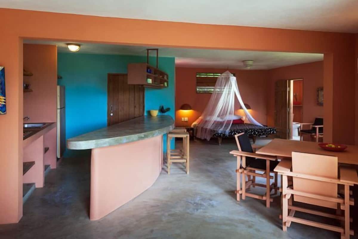A room with a curved kitchen counter, a dining area with wooden chairs, a bed with a canopy, and a small stool.