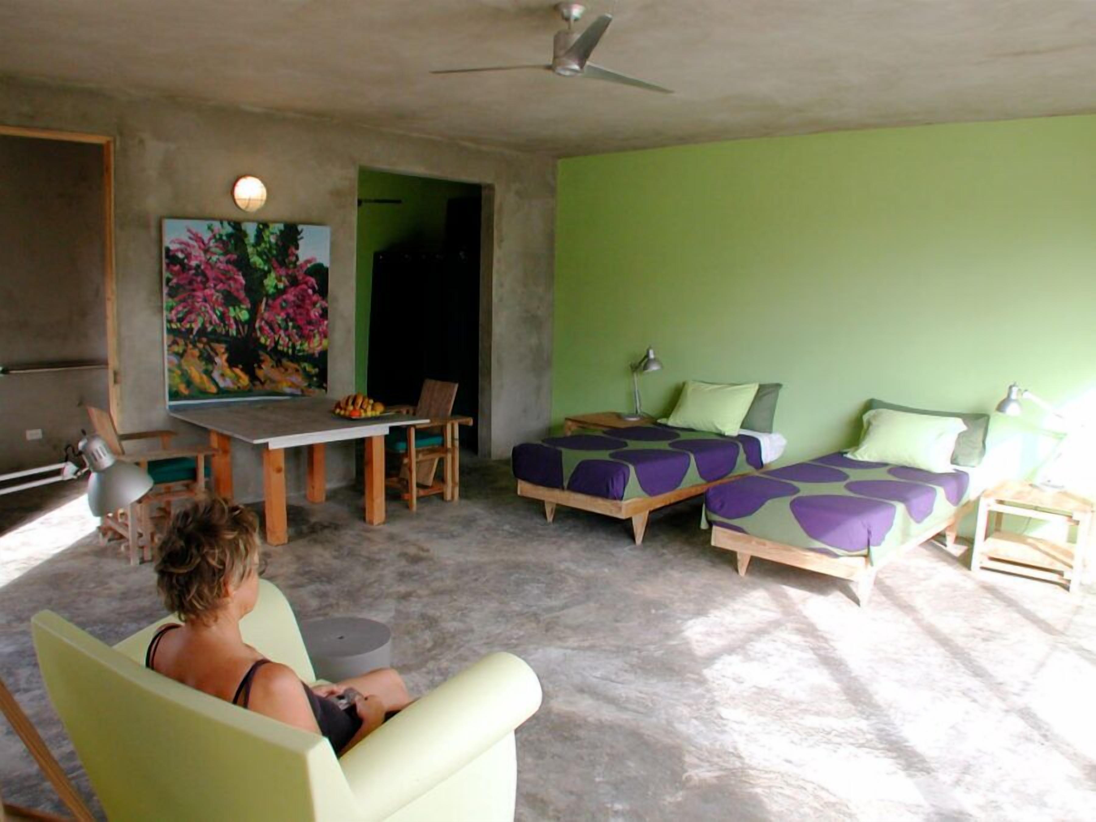 A room with a green wall, a dining table with chairs, a bed with purple bedding, and a ceiling fan.