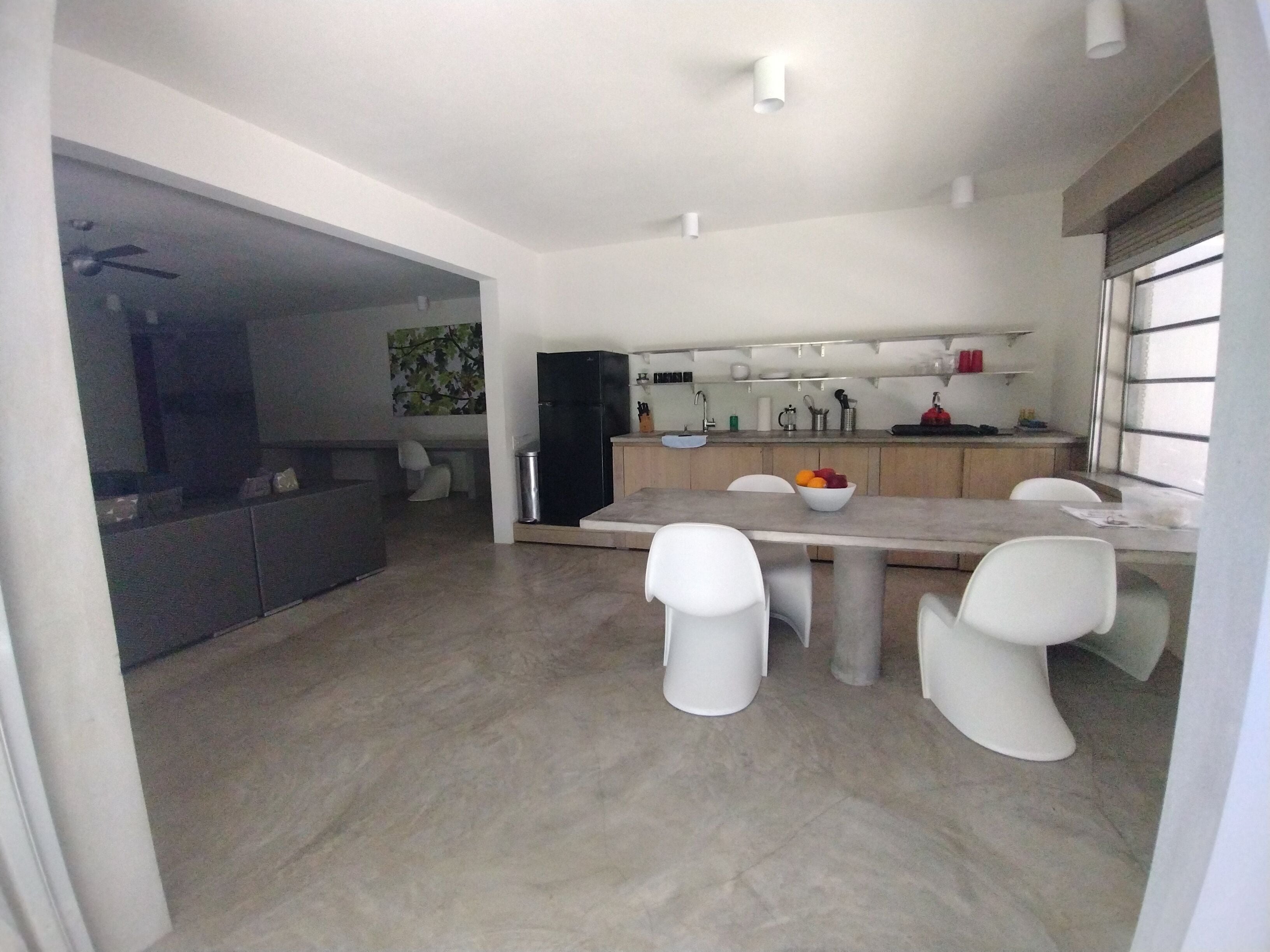 A modern kitchen with a dining area, a black refrigerator, white chairs, and a bowl of fruit on the table.