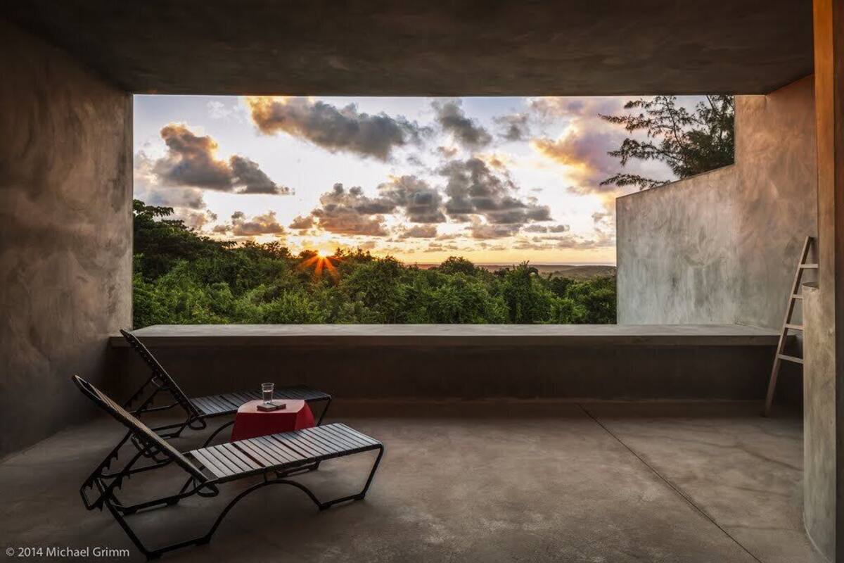 A balcony with a view of a sunset over a forest, a red table with a glass, and two lounge chairs.