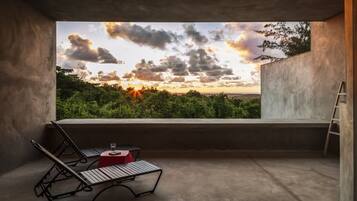 A balcony with a view of a sunset over a forest, a red table with a glass, and two lounge chairs.