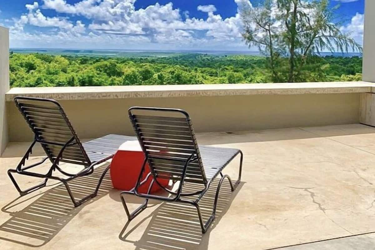 A balcony with two metal chairs and a red object, overlooking a lush landscape with trees and a clear blue sky.