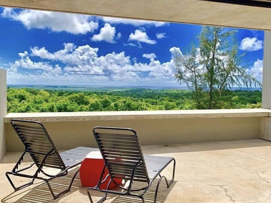 A balcony with two metal chairs and a red object, overlooking a lush landscape with trees and a clear blue sky.