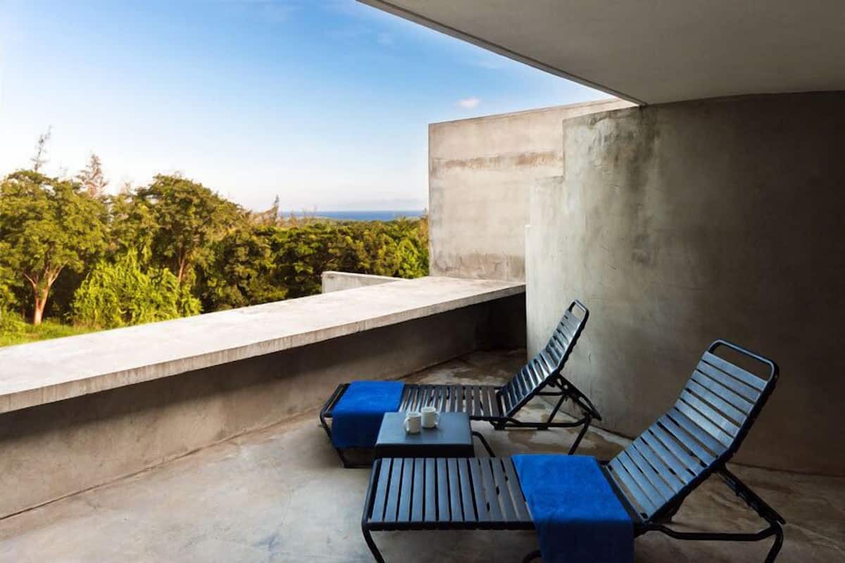 A balcony with two lounge chairs and a small table, overlooking a lush green landscape and the sea.