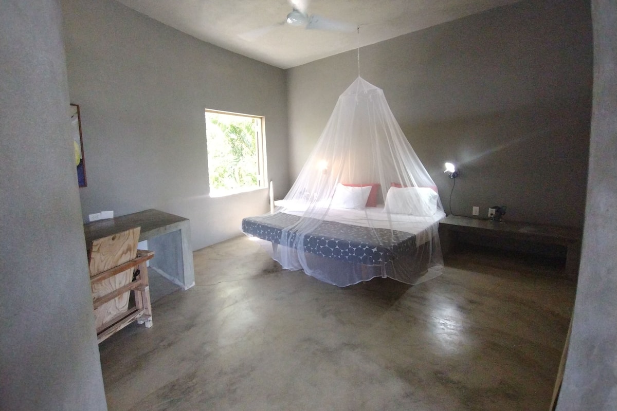 A bedroom with a bed under a mosquito net, a small desk, a window with a view, and a ceiling fan.