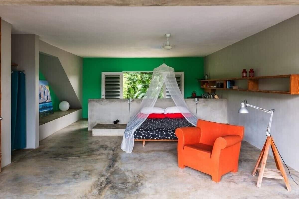 A bedroom with a bed under a canopy, a red chair, a desk lamp, and a green wall.