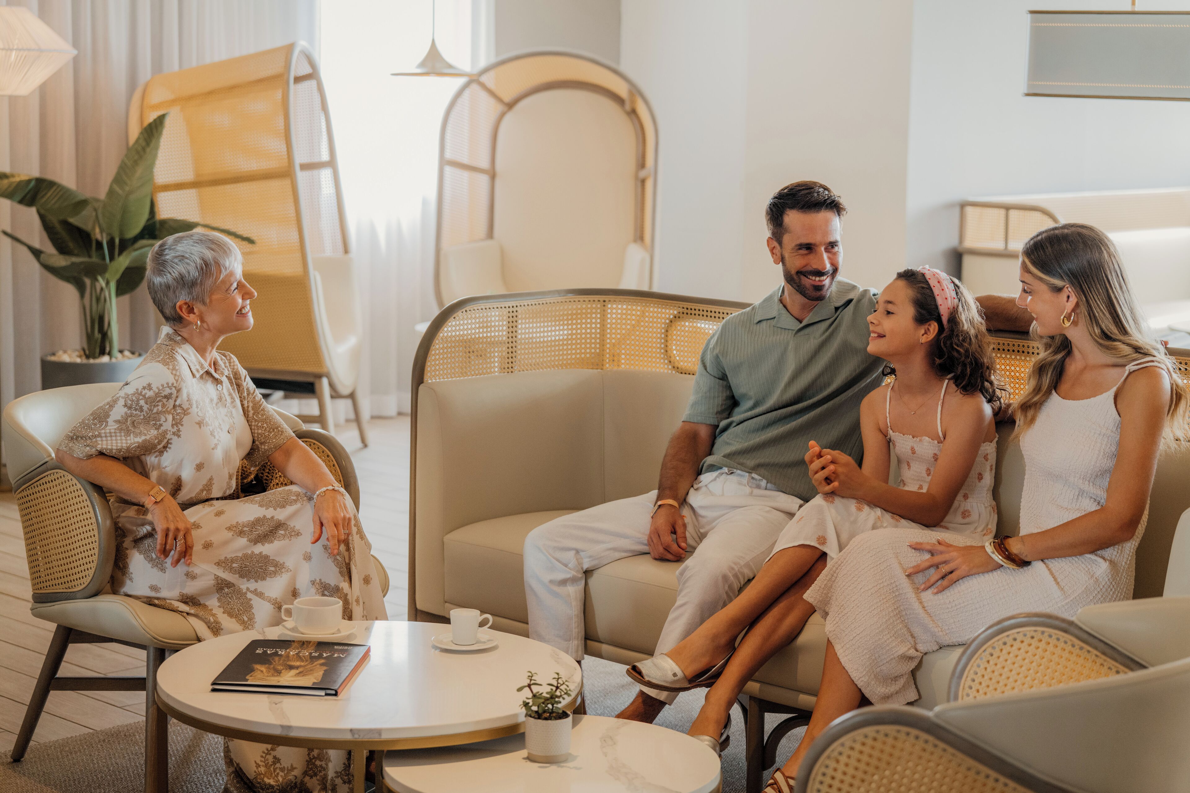 Una familia de cuatro personas sentadas alrededor de una mesa redonda en una sala de estar moderna, con un sofá, sillas y una planta en maceta.