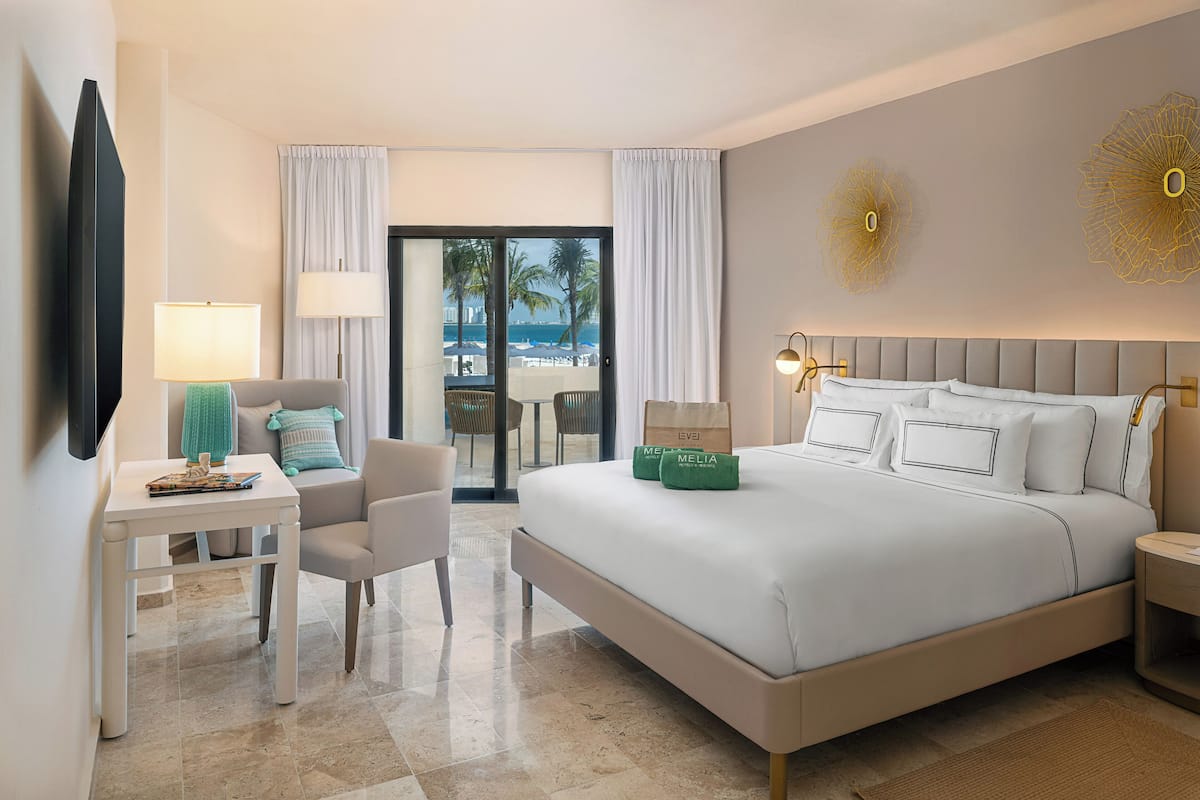 A hotel room with a large bed, a desk with a lamp, a TV, and a view of the beach through the window.
